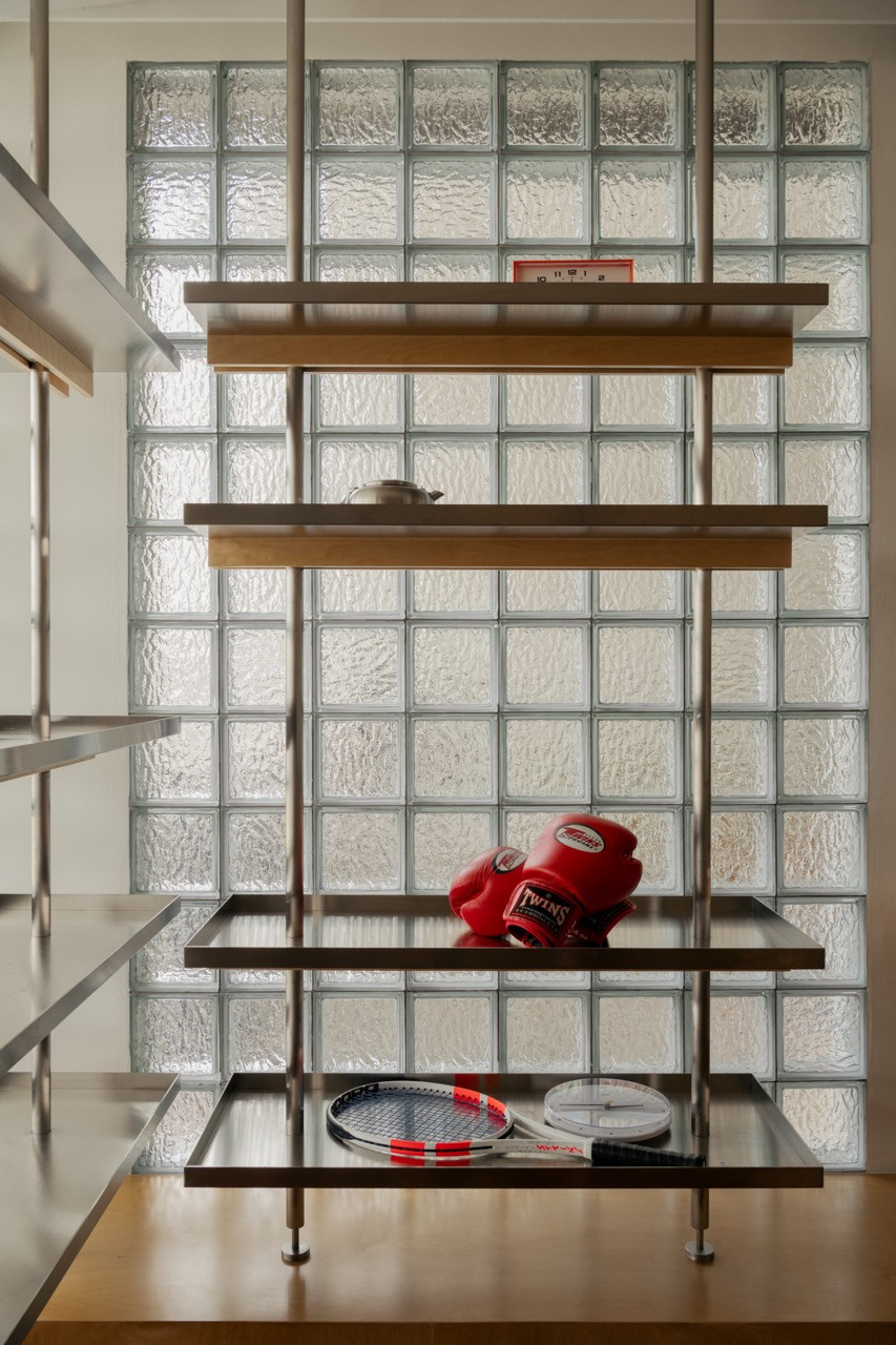 A steel and timber shelving unit set against a glass-block wall, holding sporting equipment and a clock