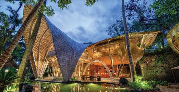 Ulaman’s reception and dining pavilion lit at dusk, its shingled bamboo roof arching over a reflecting pool at the entrance.