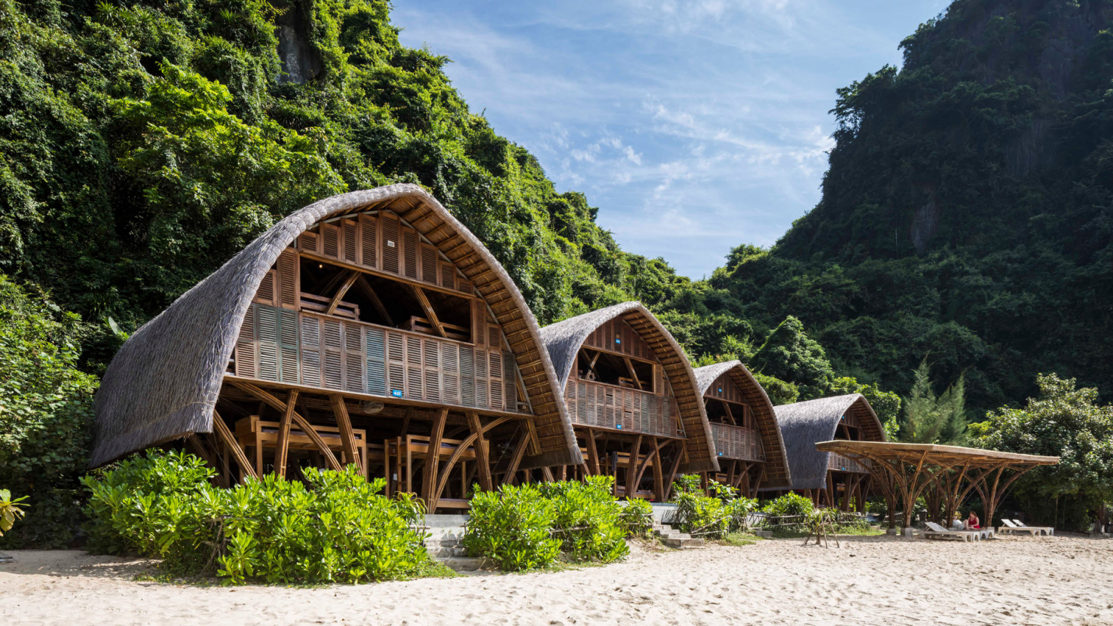 A row of guest huts at Castaway Island Resort on Cat Ba, Vietnam, their curved thatched roofs rising directly from bamboo-framed lower floors set into the beach