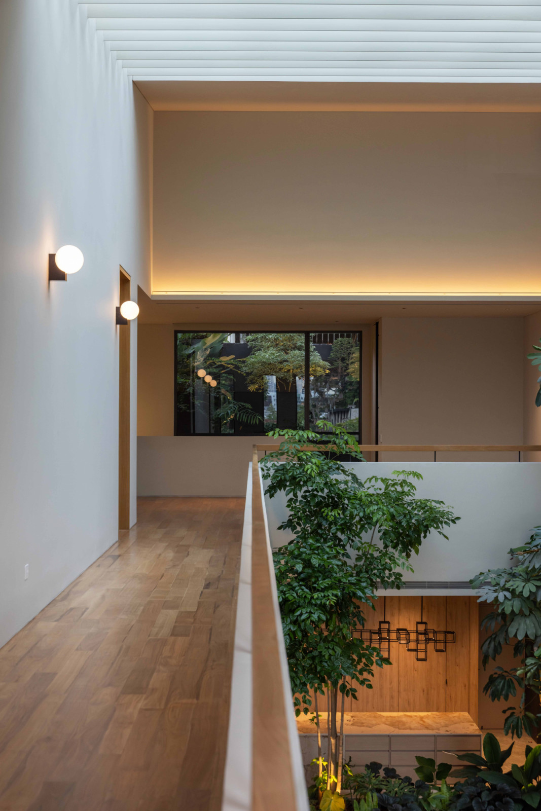 A bedroom corridor overlooks the atrium’s indoor garden, where warm cove lighting and globe sconces set a contemplative evening mood