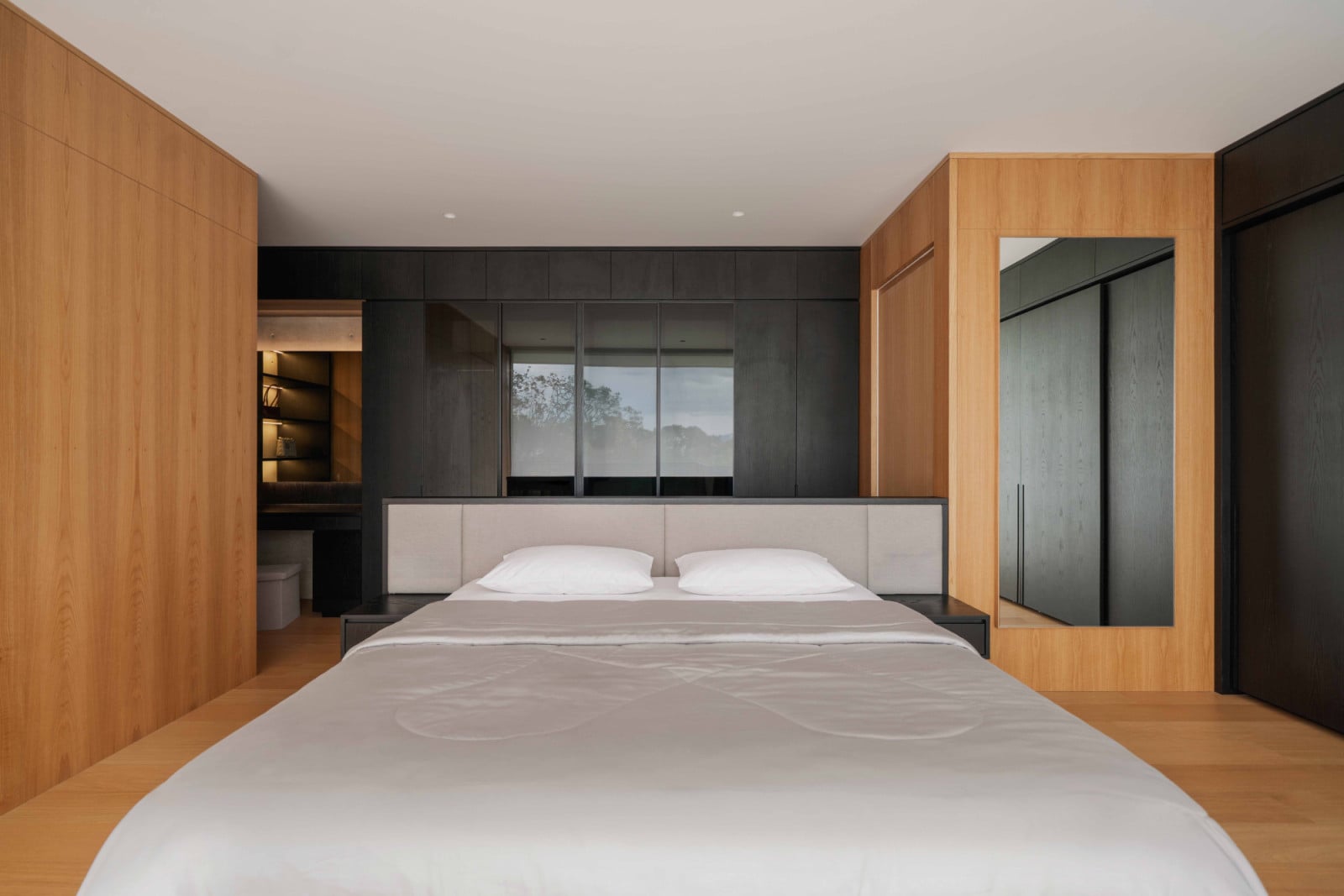 The Bandung house’s master bedroom stands behind dark, built-in joinery, with a mirrored panel doubling the light from the terrace beyond