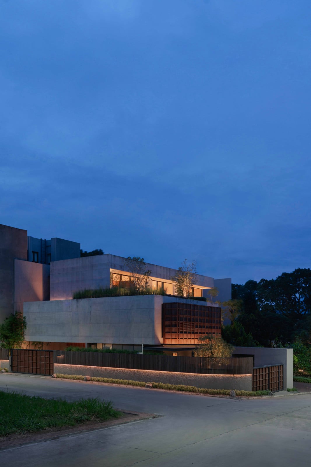 At dusk, Lift House's concrete mass and timber screen glow above its guarded street wall in Bandung