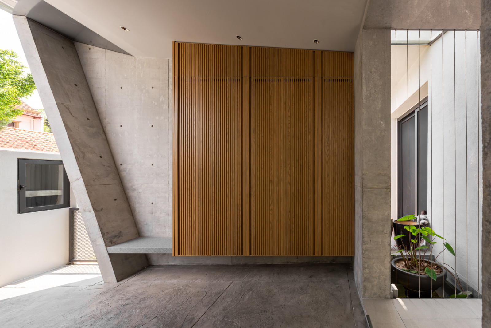 Timber slat panelling lines a covered outdoor space near the entrance, beside an angled concrete structure and a small planted courtyard