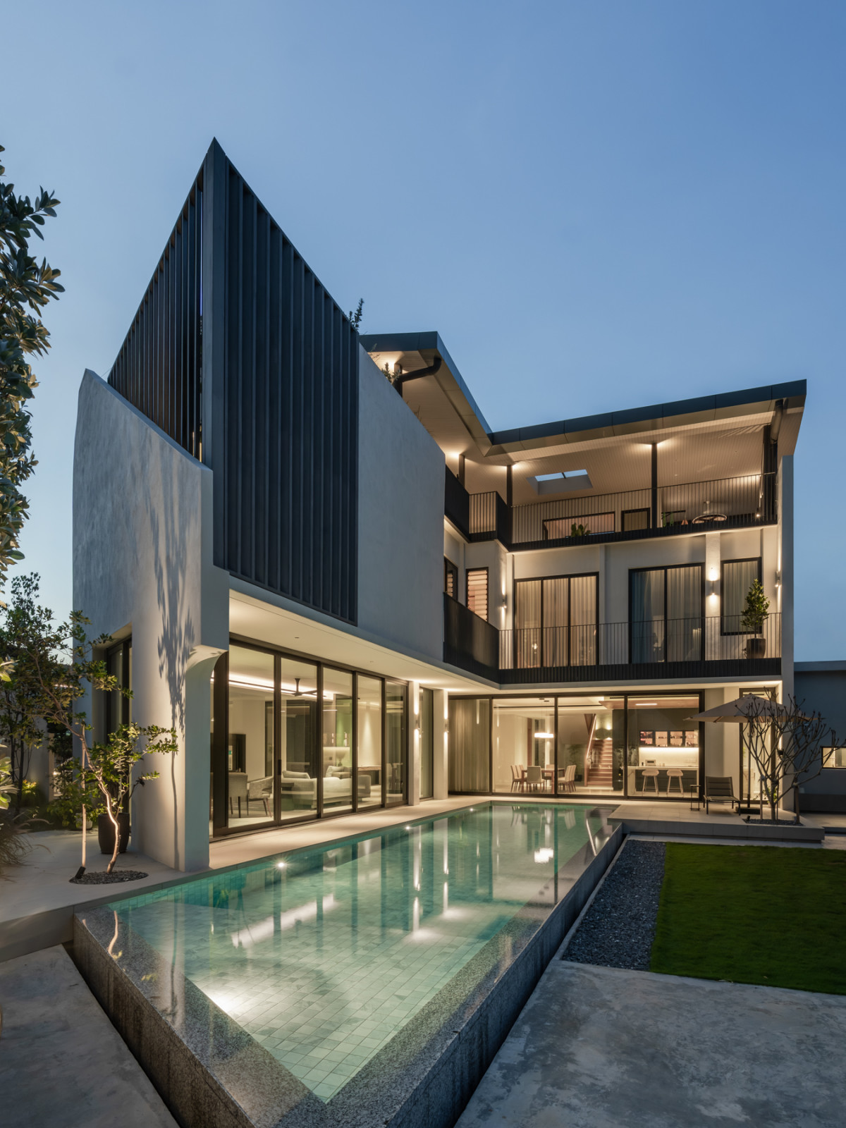 The rear elevation at dusk shows the roof terrace’s angled steel-clad form above the pool and garden