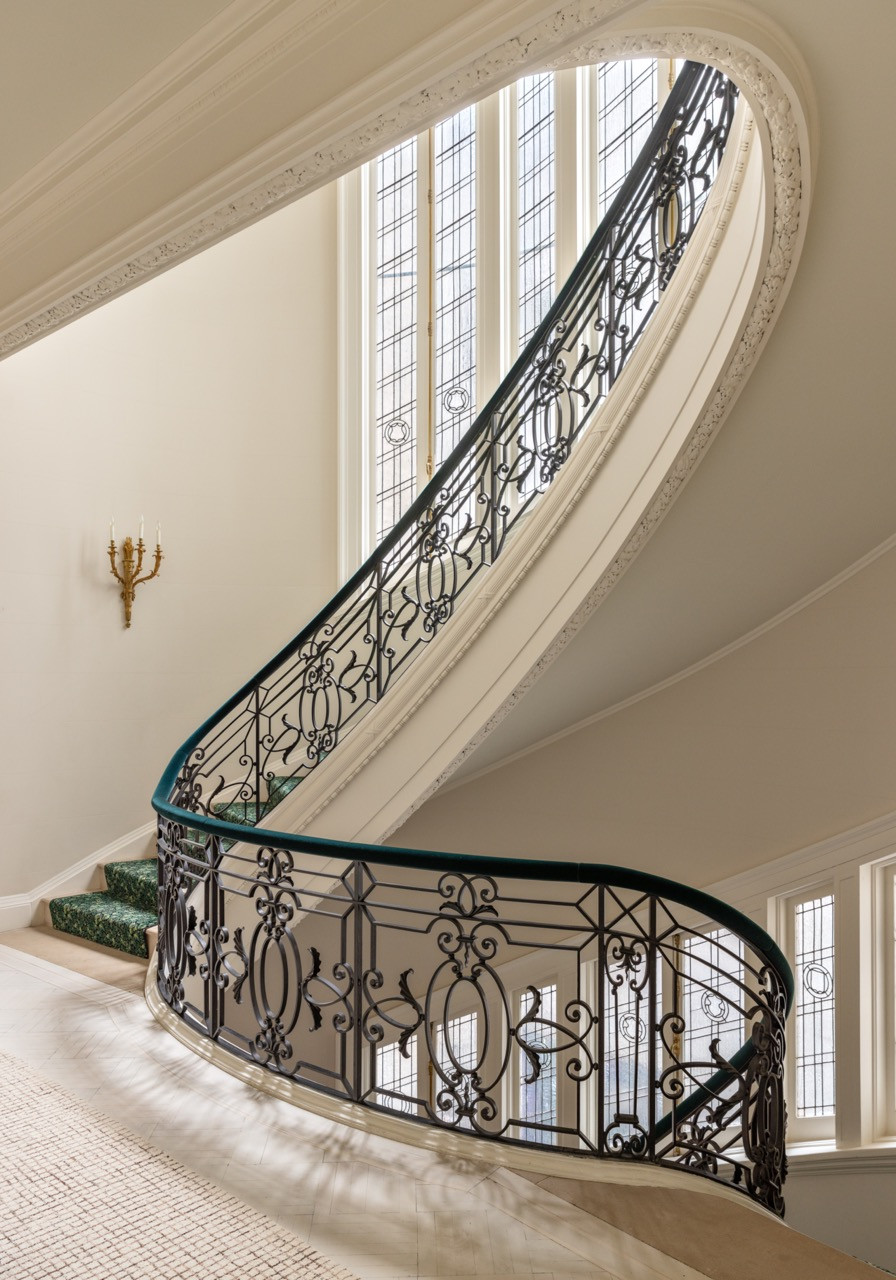 A wrought-iron balustrade curves along the mansion’s staircase, its treads carpeted in green.