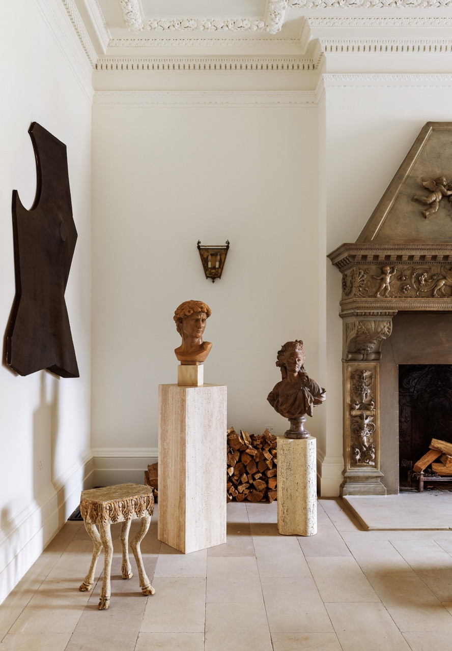 Terracotta busts on travertine pedestals stand beside the same carved stone mantel
