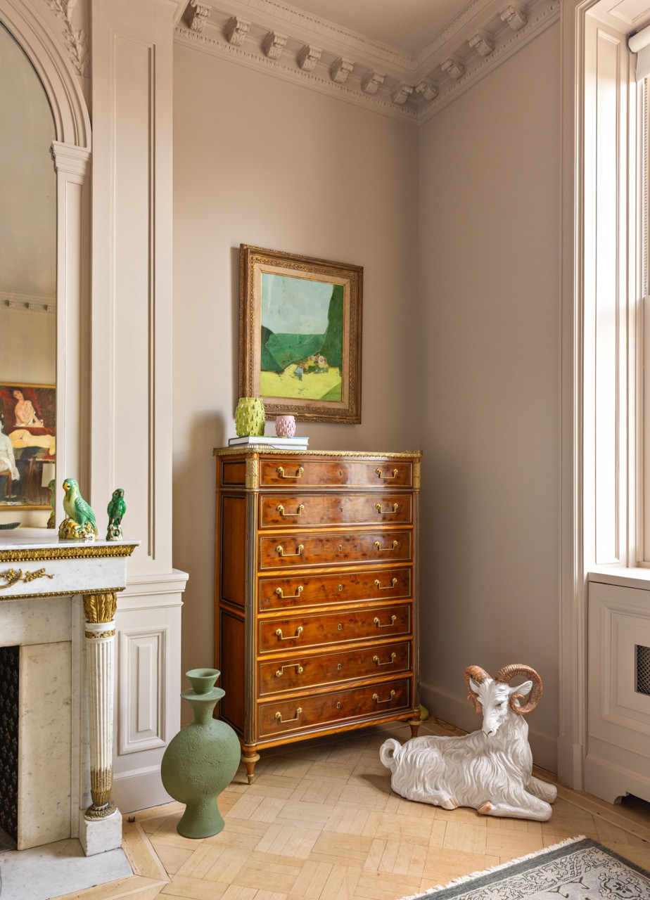 A pair of ceramic parrots and a marble mantel share a corner with an antique chest of drawers and a taxidermy ram figure