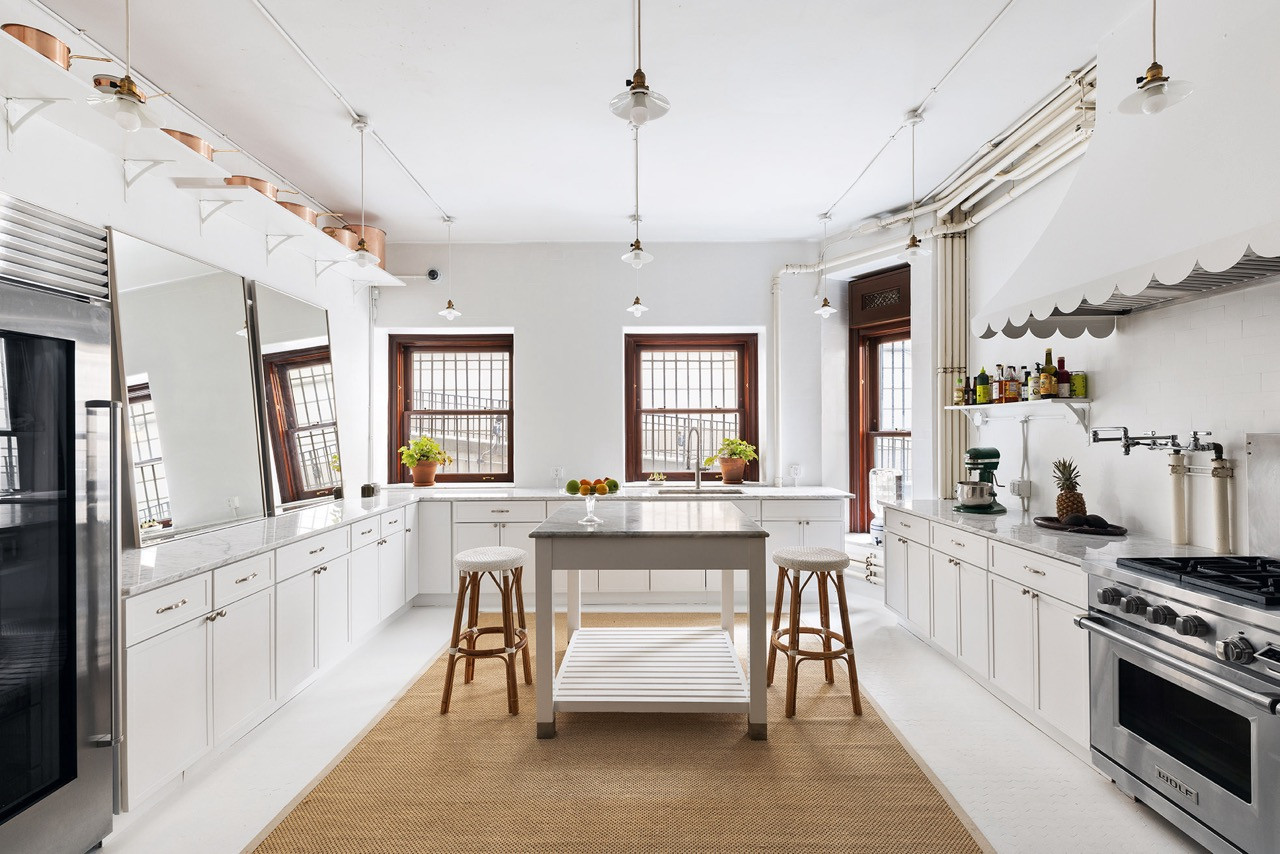 The kitchen’s white cabinetry and marble counters sit beneath a run of copper cookware shelving and a bank of factory-style pendant lights