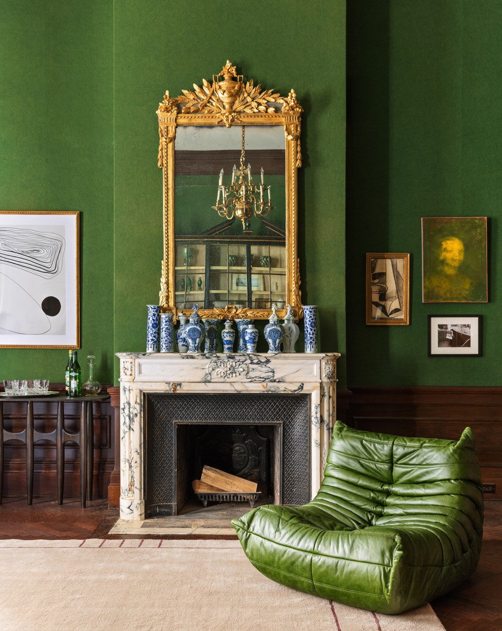 Blue-and-white porcelain vases line the marble mantel beneath a gilt mirror, with a green leather Togo chair pulled up to the fire
