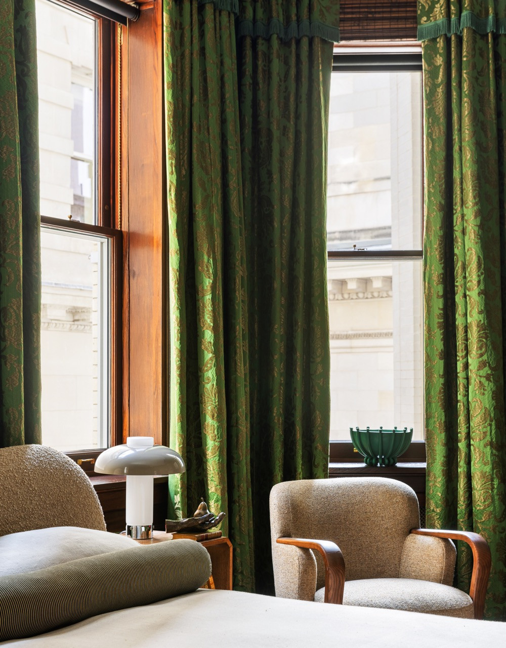 Two bouclé-upholstered armchairs flank a window framed by green silk curtains in the same emerald bedroom