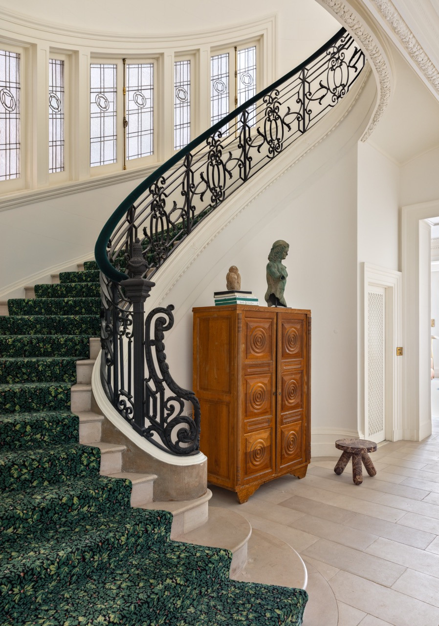 A carved wooden cabinet, topped with a bust and an owl figurine, stands beneath the staircase’s wrought-iron railing