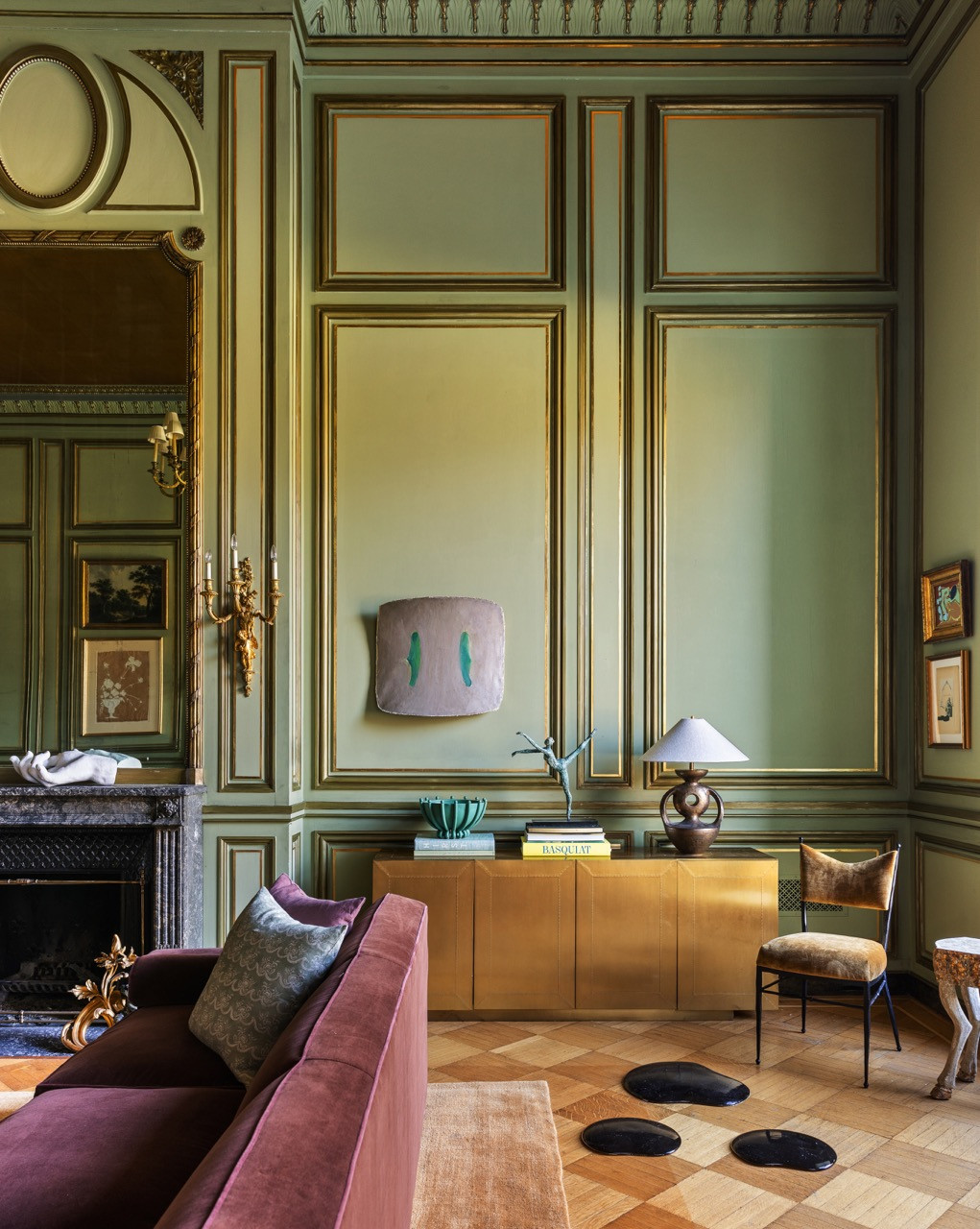 A green-panelled wall hung with an abstract painting, framed by a gilt mirror and brass sconces