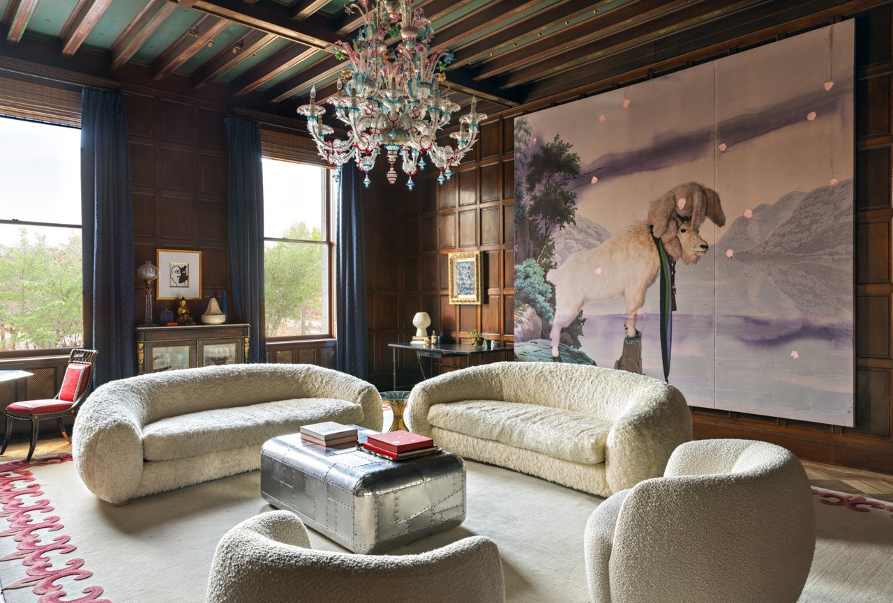 A wood-panelled sitting room holds bouclé sofas, a riveted metal trunk, and a large-scale painting of a goat, beneath a Murano glass chandelier