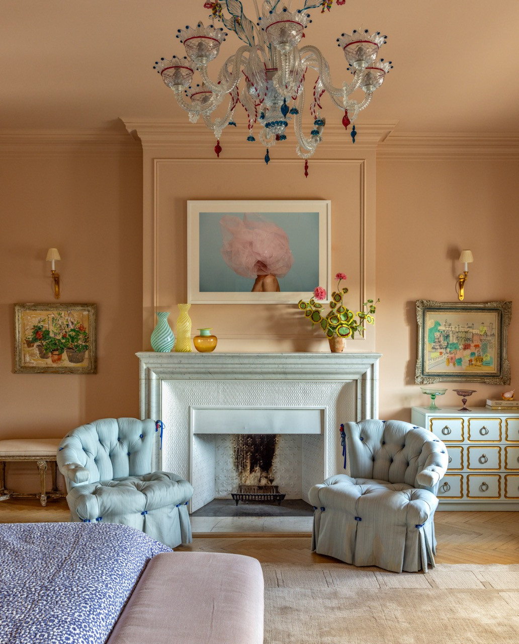 A primary bedroom in dusty pink, its tufted chairs flanking a marble mantel beneath a Murano chandelier and a framed photograph