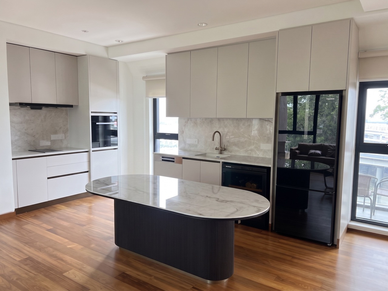 A pale kitchen with an oval marble-topped island, integrated appliances and a marble splashback opens onto a balcony through glazed doors