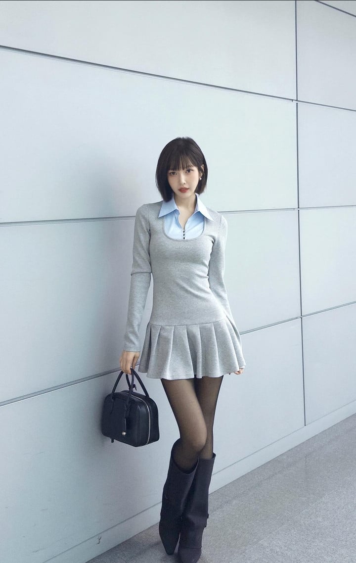 A woman wears a grey long-sleeve dress with a pleated skirt, a light blue collared top, black boots, and a handbag.