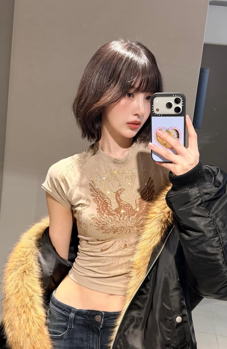 An individual captures a mirror selfie, featuring a sparkling top with an avian motif and a fur-trimmed bomber jacket.