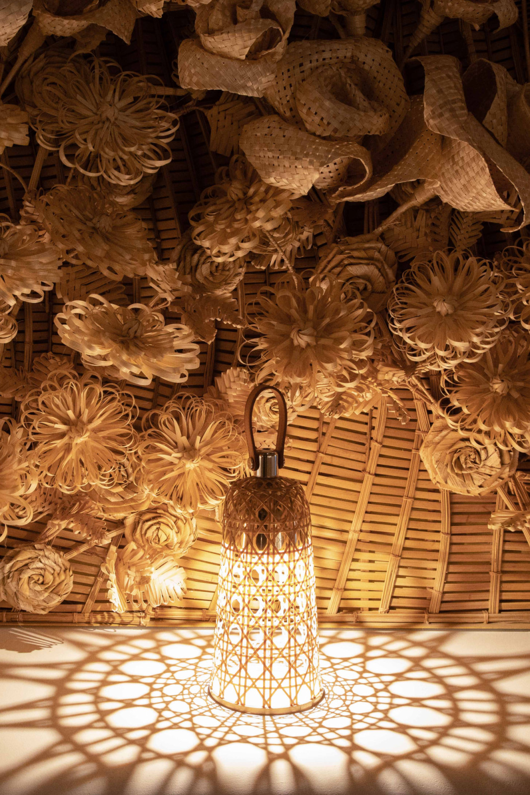 A Dior Maison Corolle lamp in woven bamboo casting its intricate geometric shadow onto the floor, set against the woven installation by Thai artists Korakot Aromdee and Vasana Saima