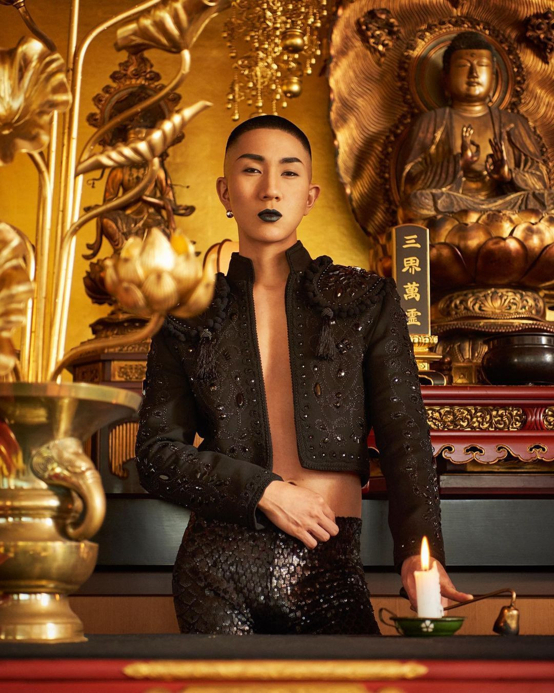 A person showcases dramatic makeup and an embellished black outfit against a backdrop of golden Buddha statues.