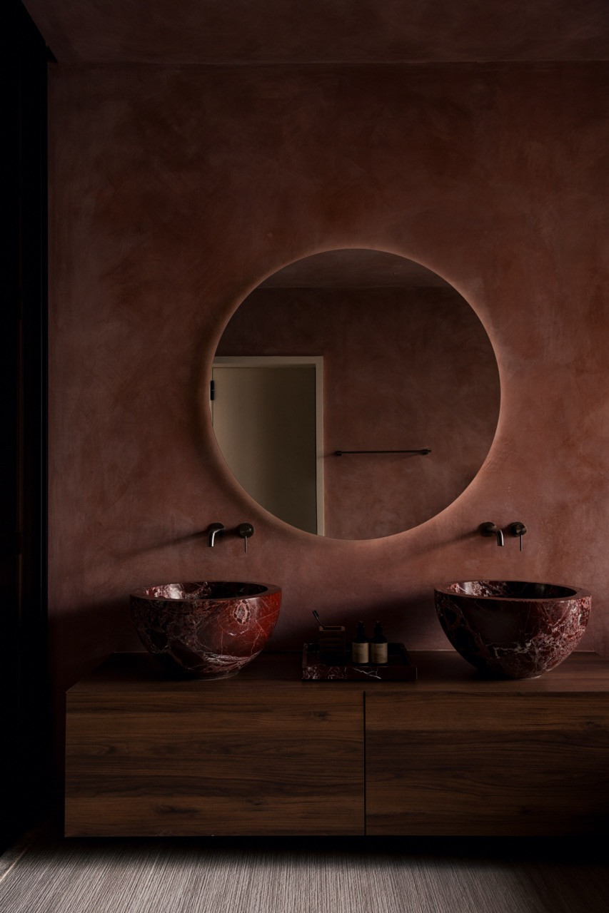 Twin vessel basins in veined red marble, set into a teak vanity beneath a single backlit mirror