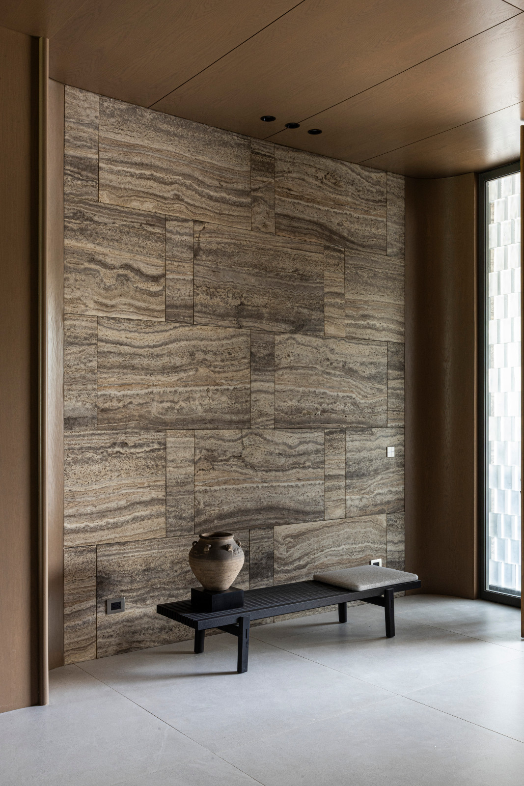 Honed Iranian travertine flows from the exterior into the foyer, where stained ash wood veneer wraps the ceiling and curved corners; a Home Hotel bench by Poliform, designed by Jean-Marie Massaud, completes the space