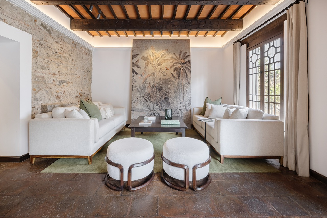 In the sitting room, an exposed stone wall stands opposite a mural depicting palms and birds, with an olive-green rug underfoot