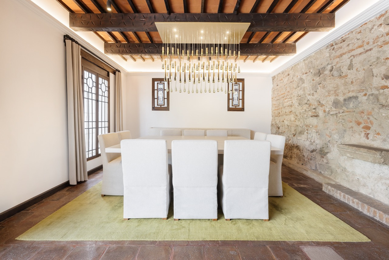 In the formal dining room, a chandelier of glass droplets hangs beneath carved beams, next to a wall of exposed stone and brick