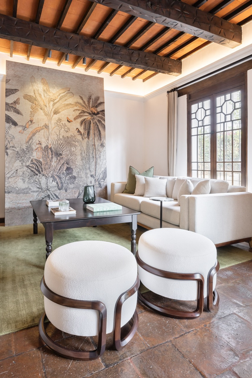 In the living room, a large-scale botanical mural hangs against carved ceiling beams, with custom upholstered ottomans by local makers