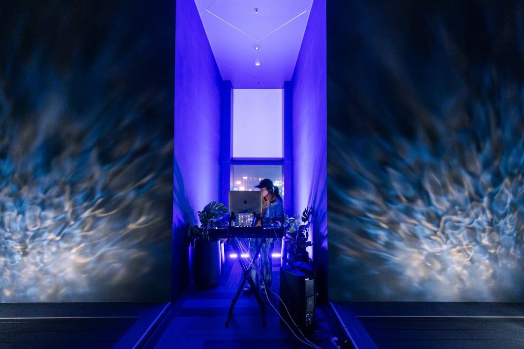A DJ performs a set at a modern bar, surrounded by ambient blue lighting and city views.