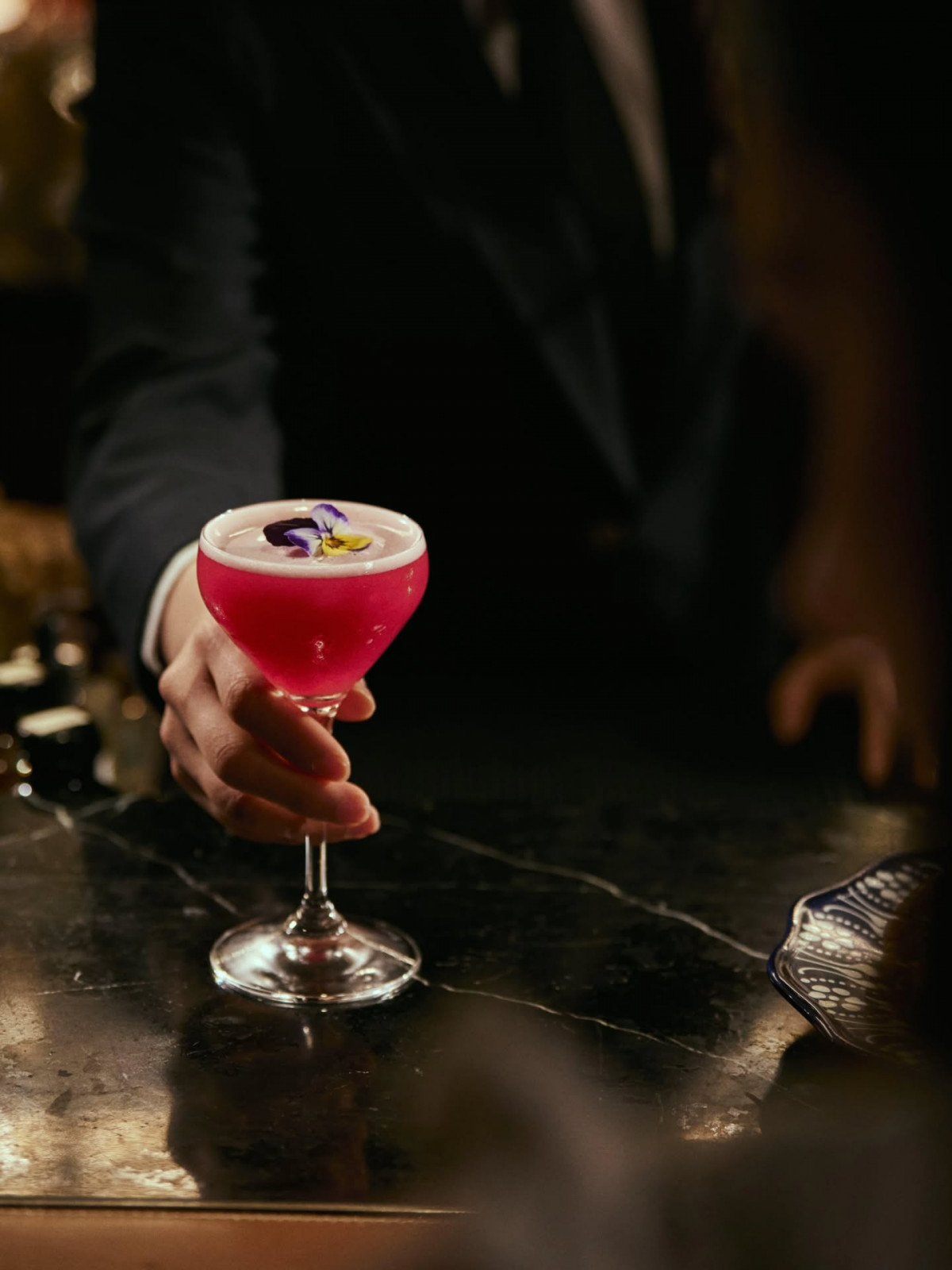 A bartender presents a vibrant pink cocktail garnished with an edible flower in a coupe glass.