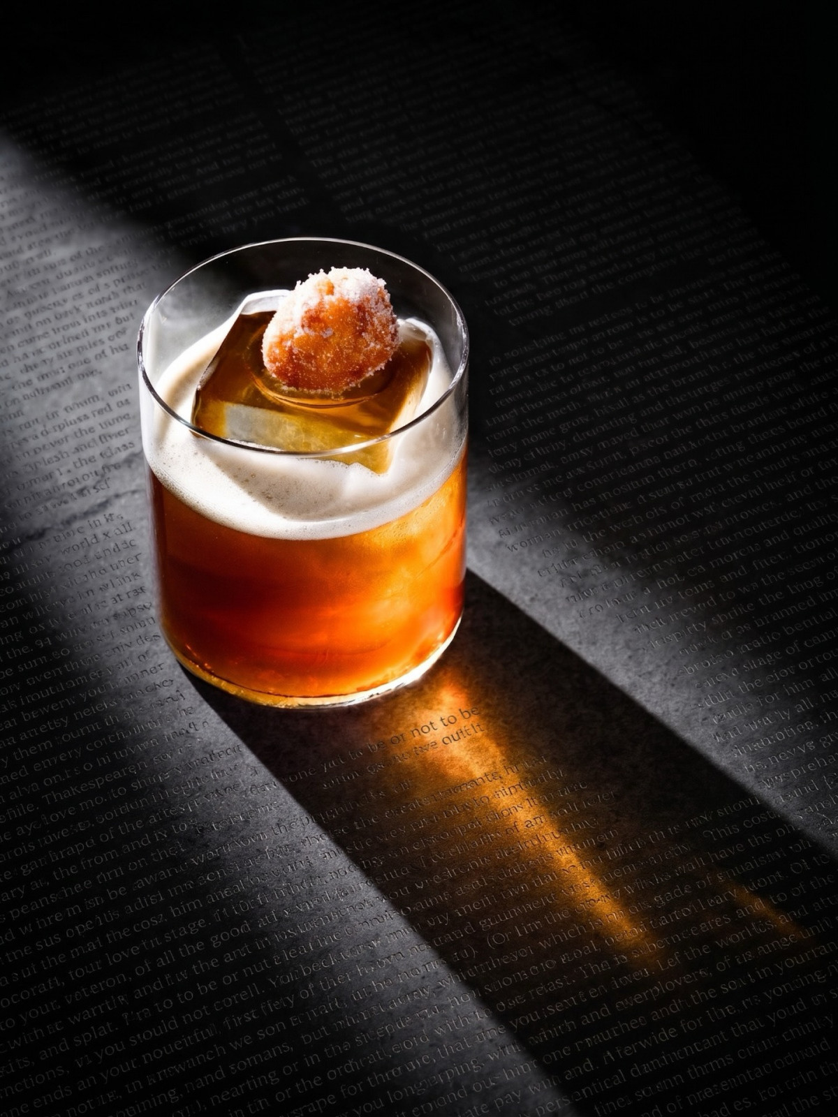 A richly colored cocktail served in a rocks glass, topped with a large ice cube and a sugar-dusted pastry garnish.