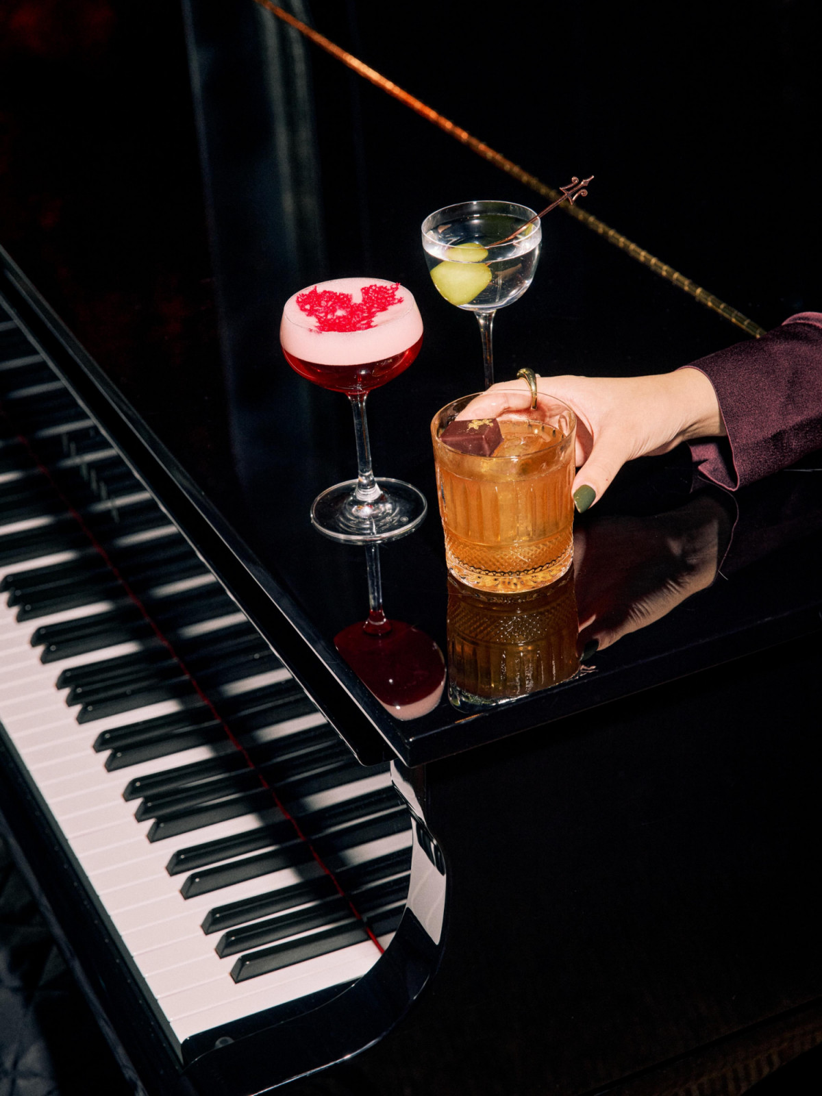Three different cocktails rest on a grand piano's polished surface, with a hand reaching for one.