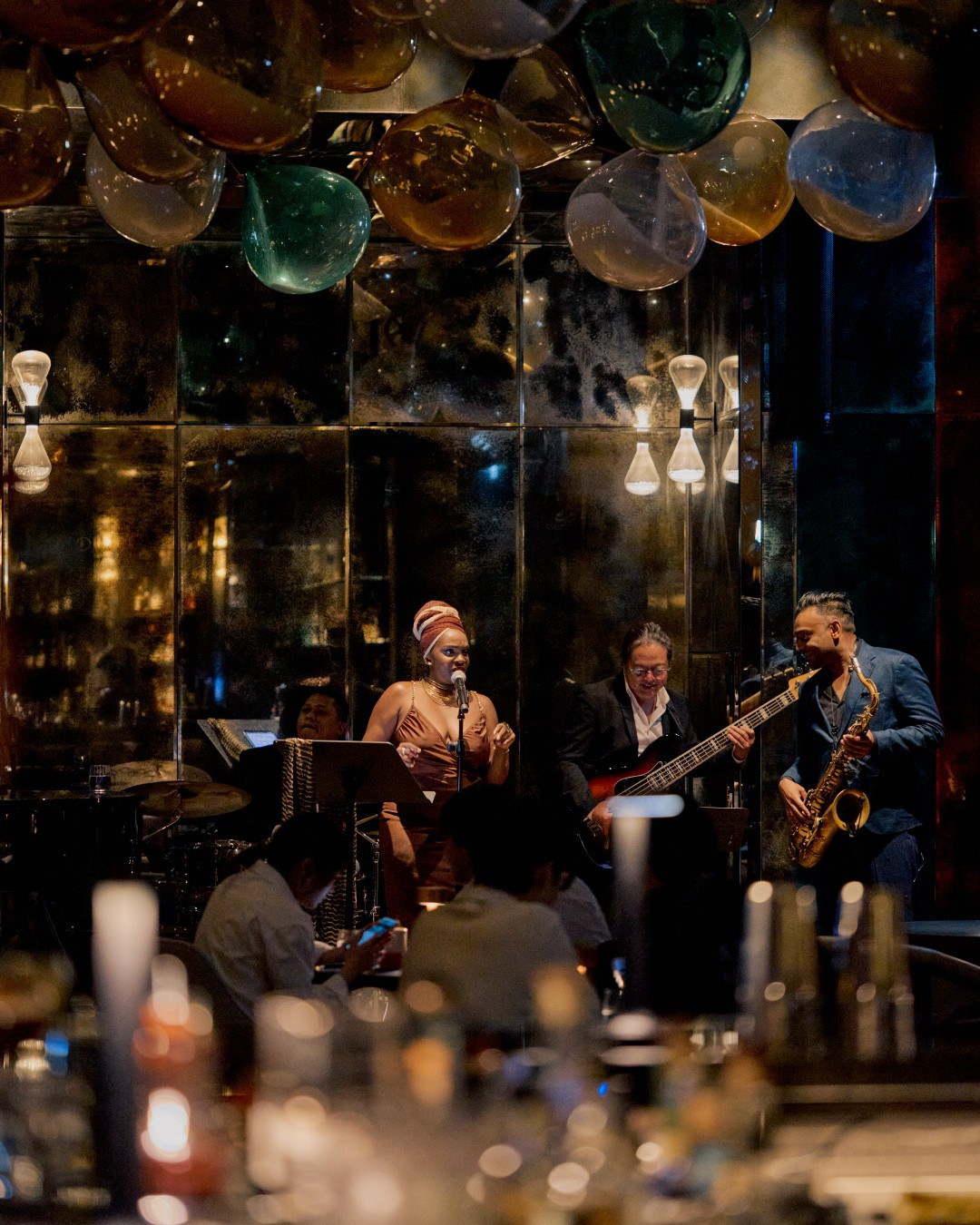 A live band performs, with a vocalist, bassist, and saxophonist entertaining guests at a bar.