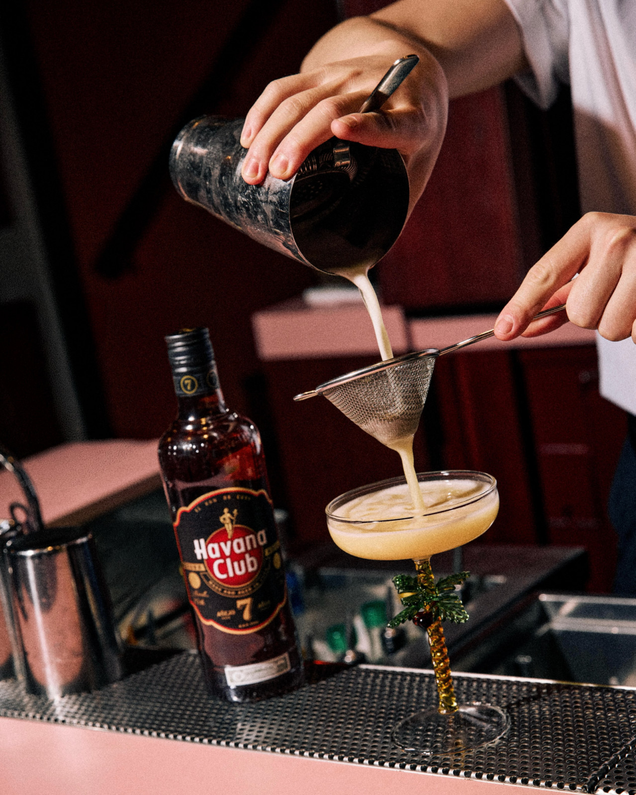 A bartender strains a Havana Club 7 rum cocktail into a glass with a palm tree stem.