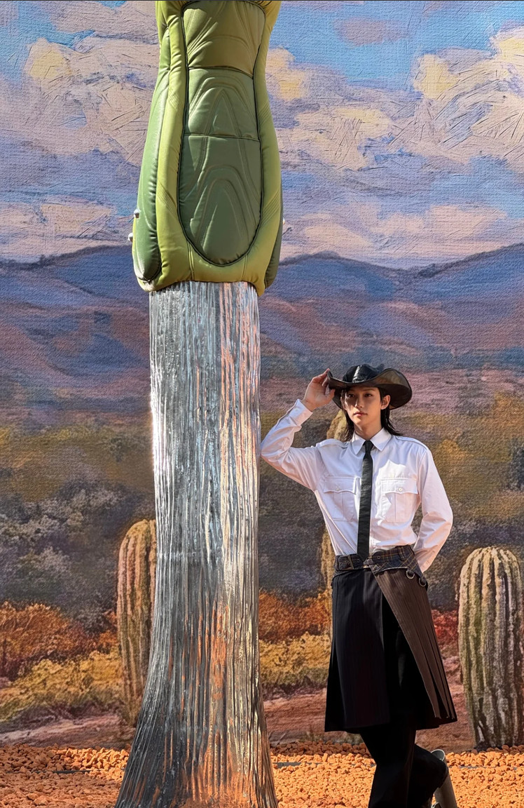 K-pop artist Felix, outfitted in a cowboy hat and a black skirt ensemble, poses against a painted desert landscape.