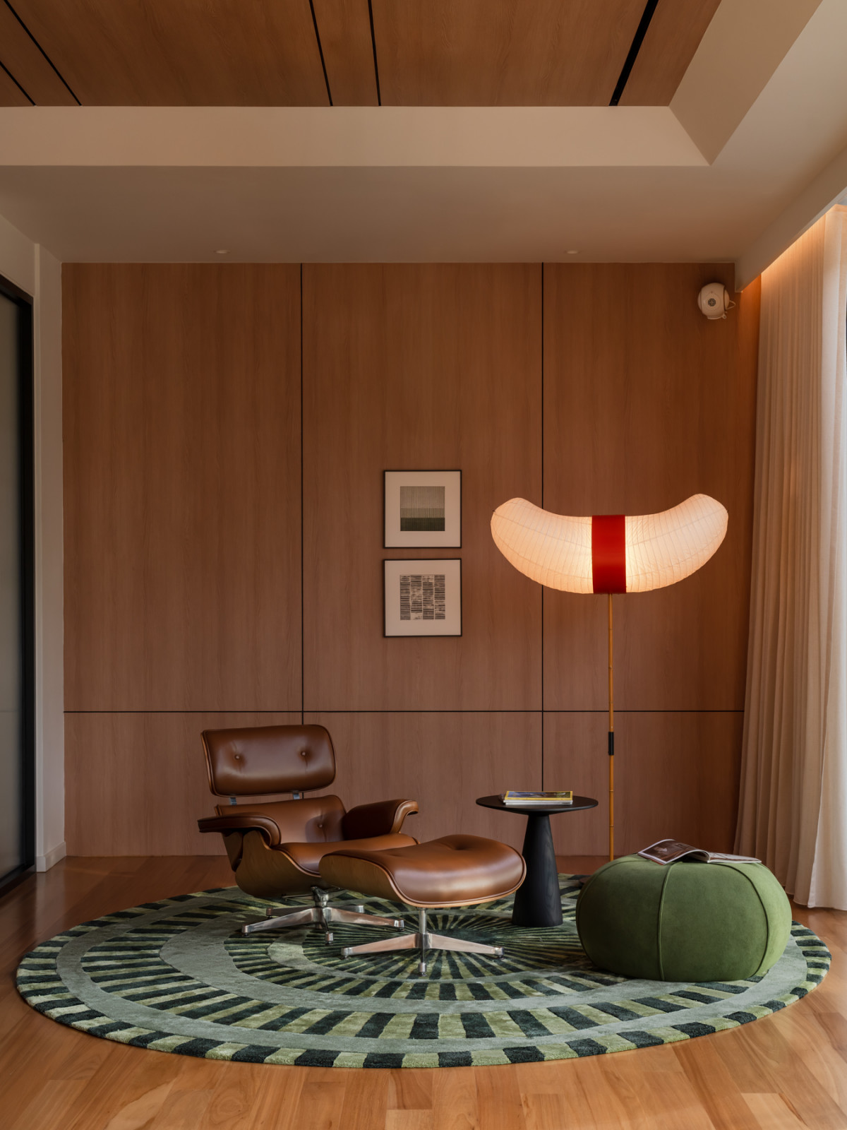 An Eames lounge chair and ottoman sit on a circular patterned rug, lit by a curved paper floor lamp against timber panelling
