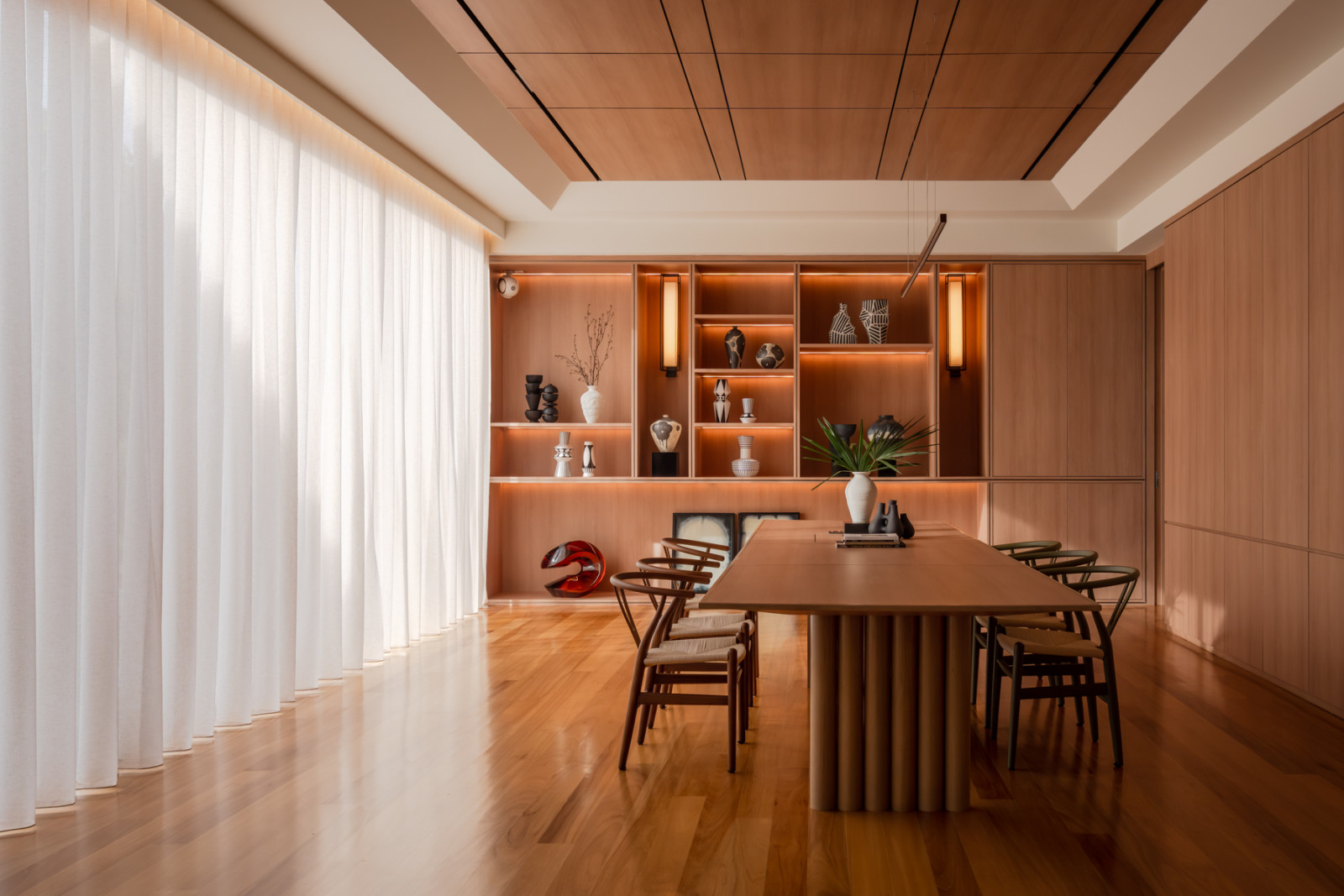 Backlit display shelving holds a collection of ceramics and vases behind the dining table, framed by sheer curtains