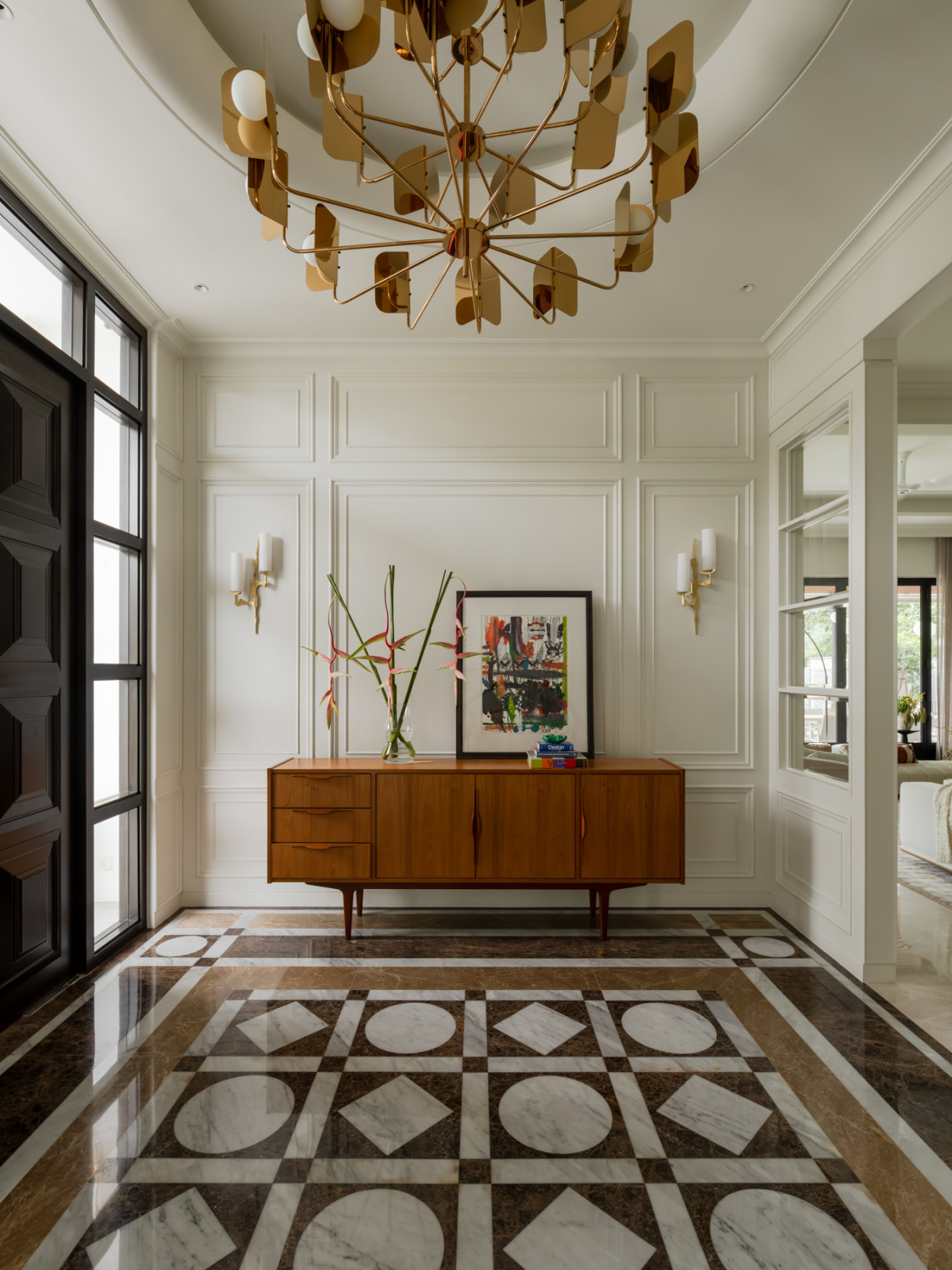 A sculptural gold chandelier hangs above the vintage teak sideboard, set against the patterned marble entrance floor
