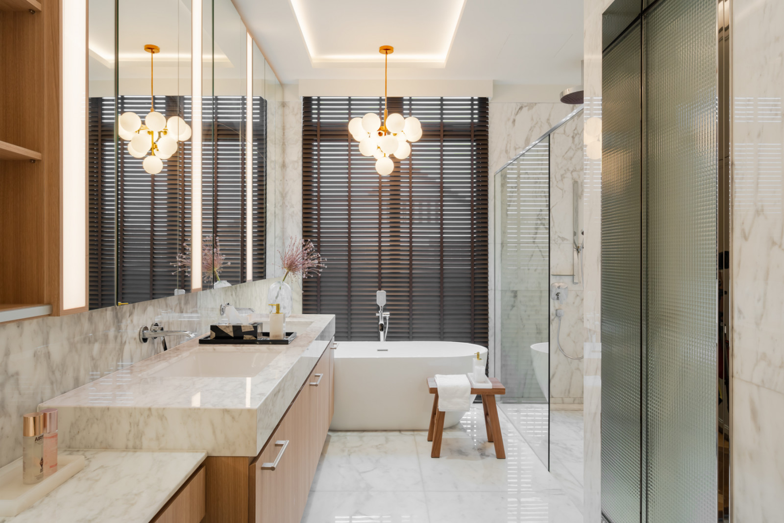 A freestanding tub and double marble vanity are lit by a globe pendant, with the shower enclosed in ribbed glass beyond