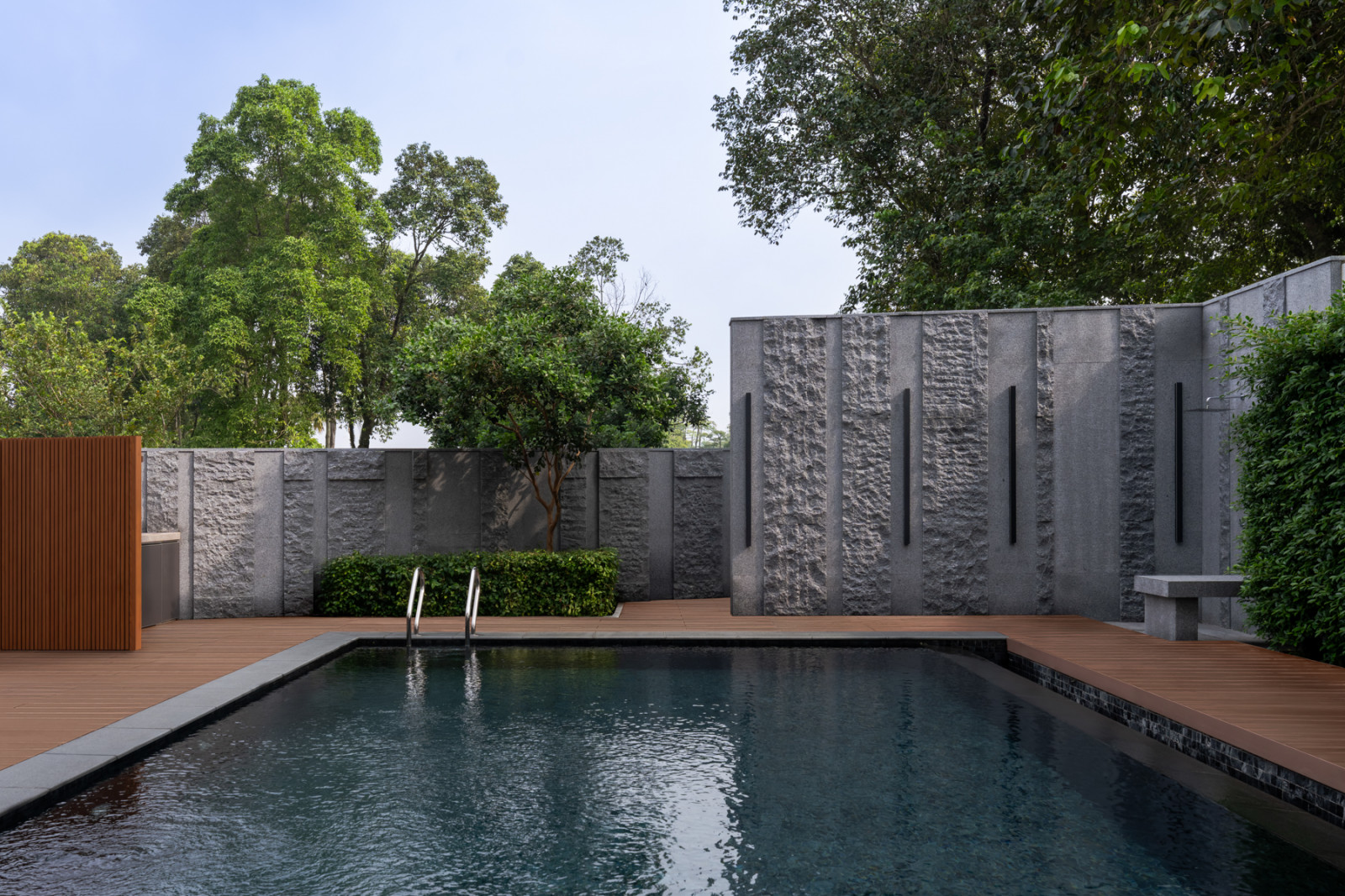 A textured stone wall with vertical light fittings borders the pool, with an outdoor shower set against a timber deck
