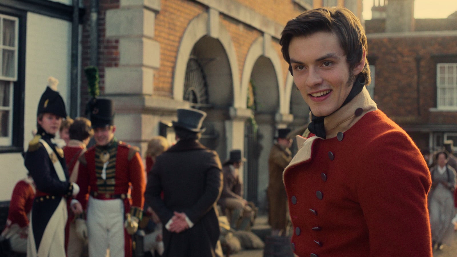 A young man in a scarlet military coat smiles in a bustling scene reminiscent of a Regency-era town.