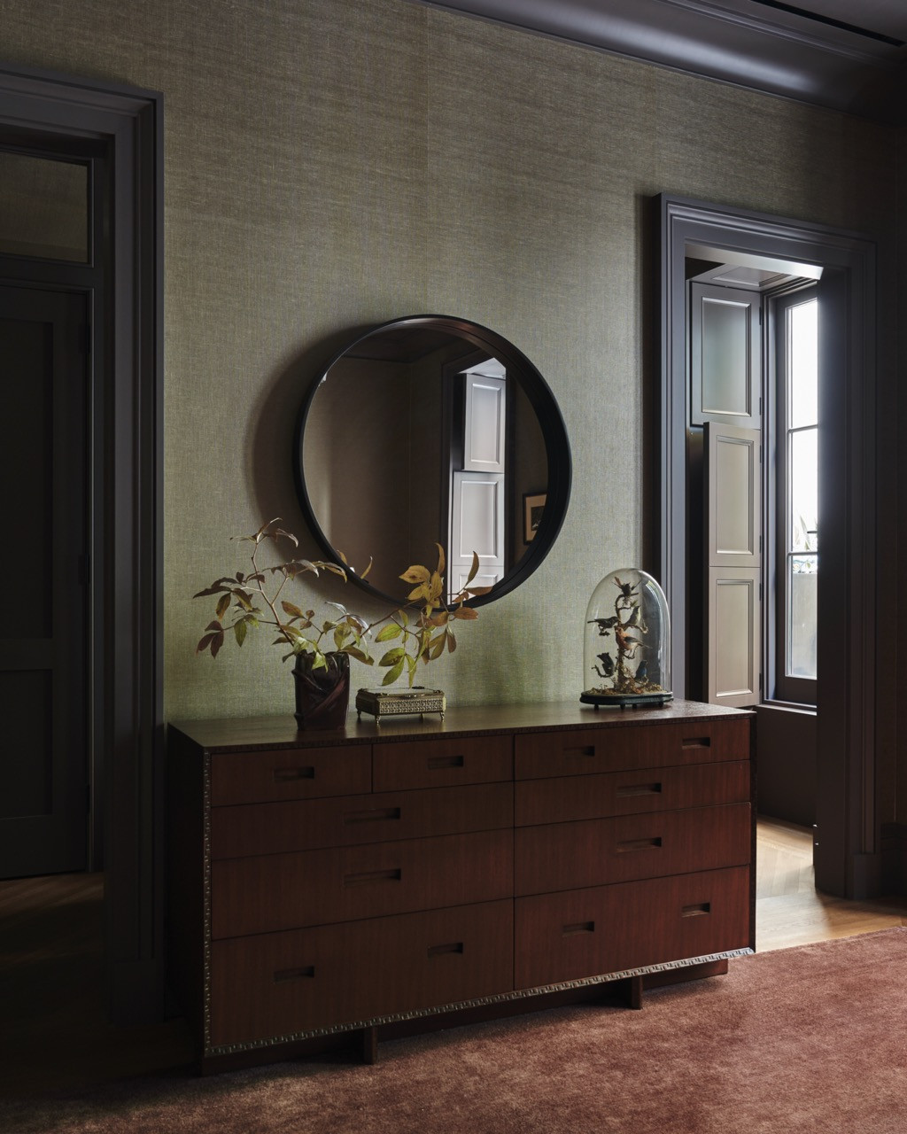 A dresser and round mirror in a grasscloth-lined room, with a glass-domed curiosity displayed alongside a branch arrangement