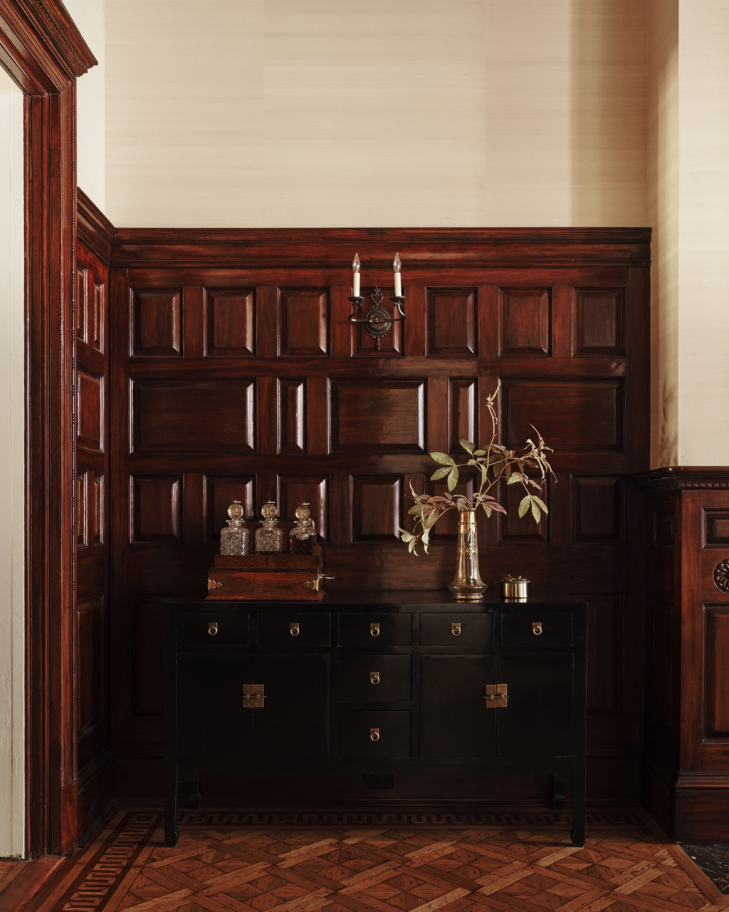 Dark wood paneling frames a black lacquered cabinet, a pair of sconces, and glass decanters — a detail consistent with the historic dining room’s restored woodwork