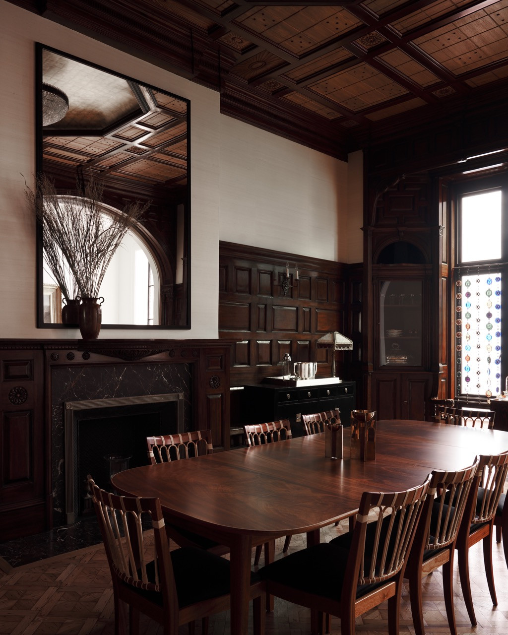 The restored dining room, with its dark panelling, coffered ceiling, and marble fireplace, original to Stanford White’s 1879 design, has a beaded glass curtain hanging at the window beyond.