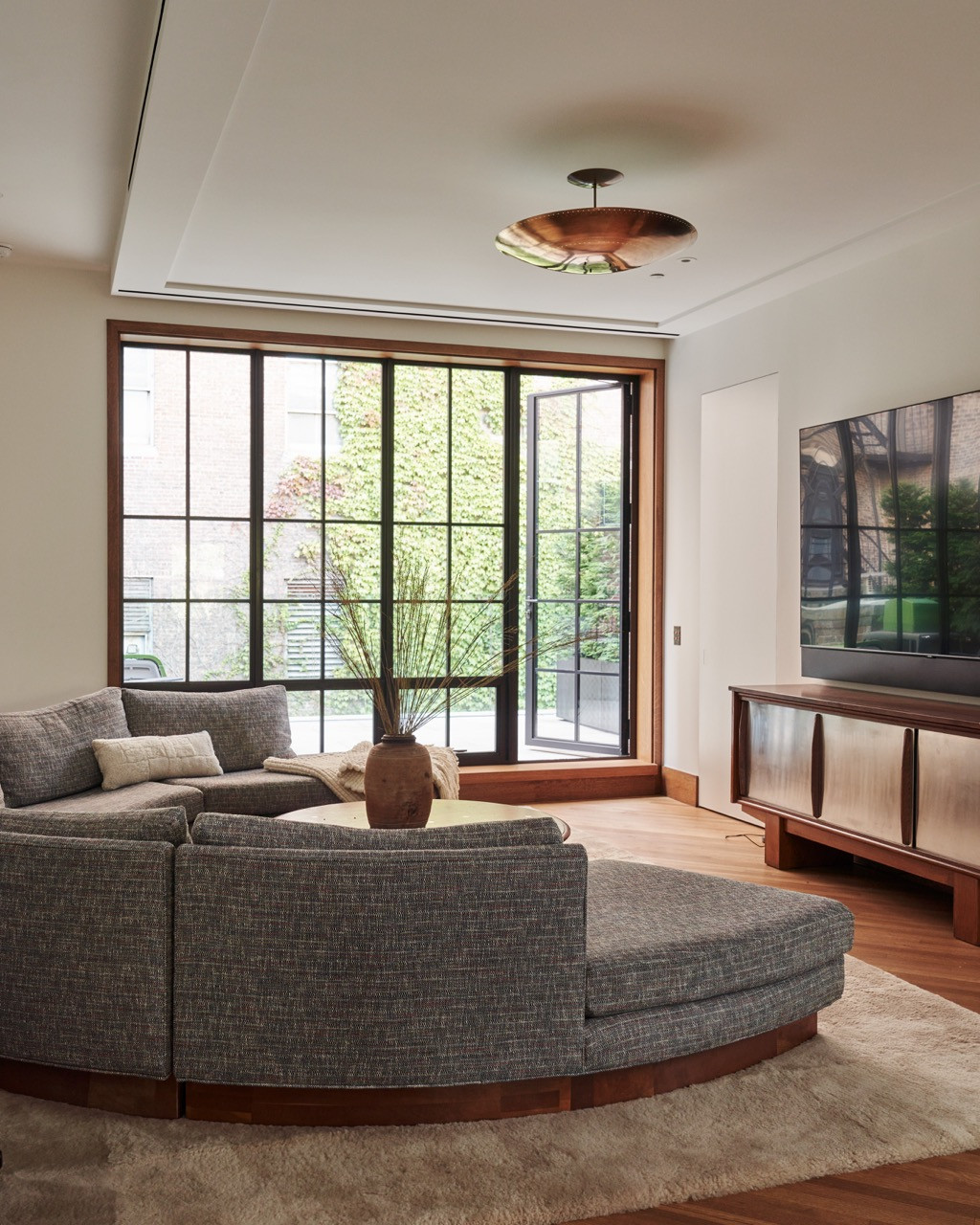 A family room with a curved sectional sofa facing a tall steel-framed door onto the ivy-covered rear garden