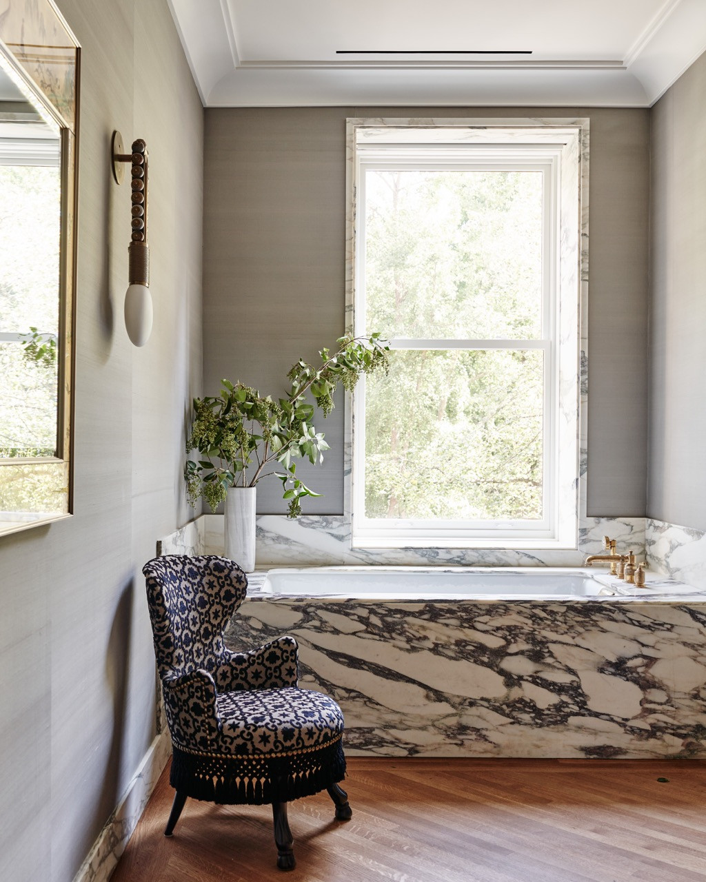 A marble-clad bathtub set beneath a tall window, with a patterned wingback chair positioned alongside