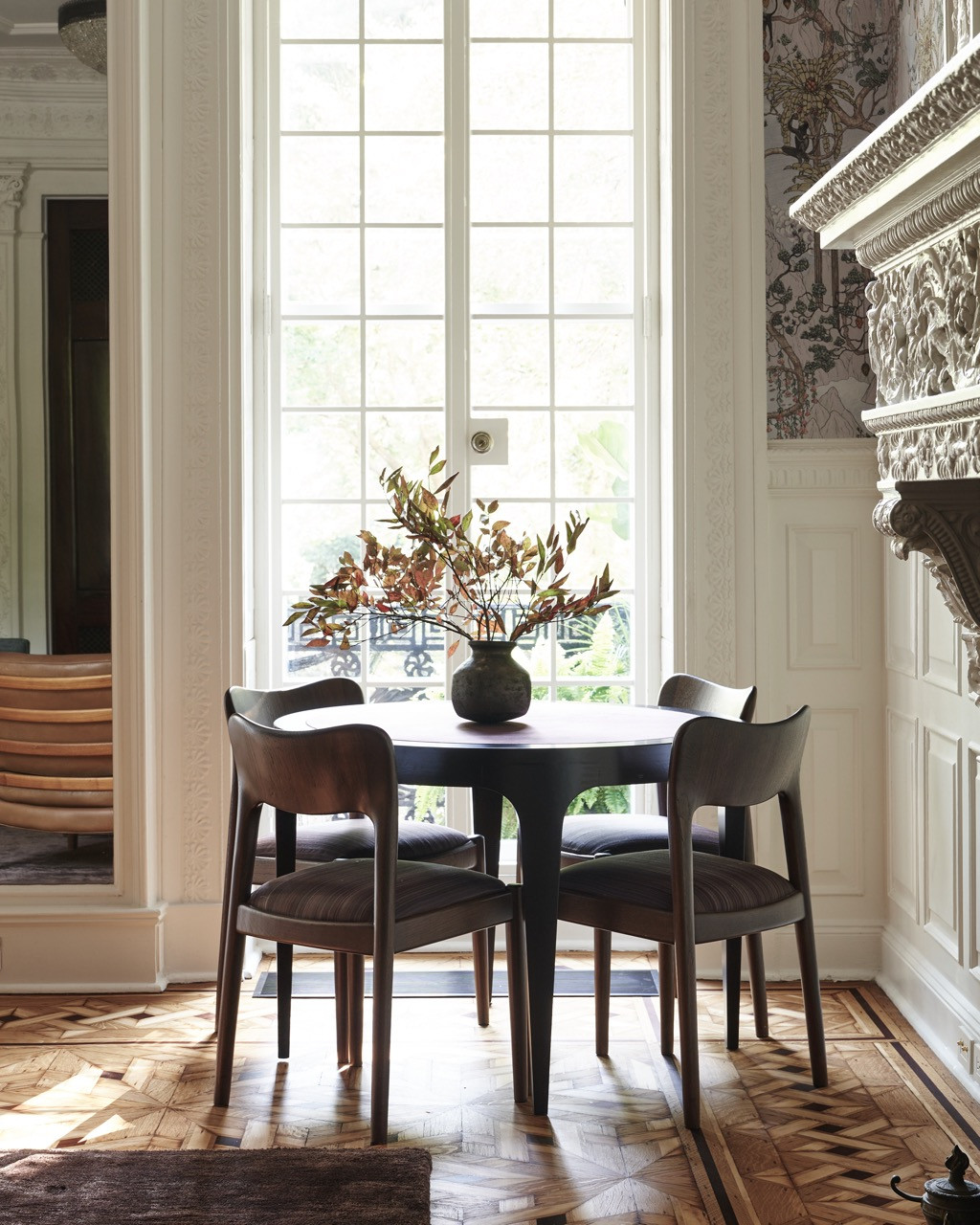 A round dining table set beneath tall windows, framed by the carved mantel and Wiseman wallpaper