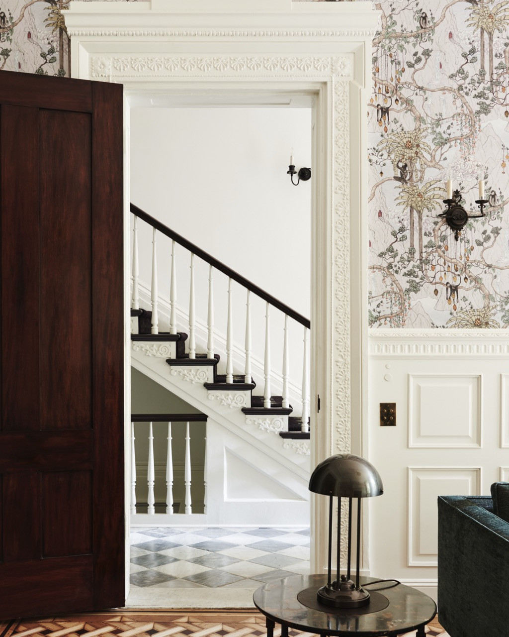 The same wallpapered room, seen through to a staircase with detailed millwork and a checkerboard marble floor