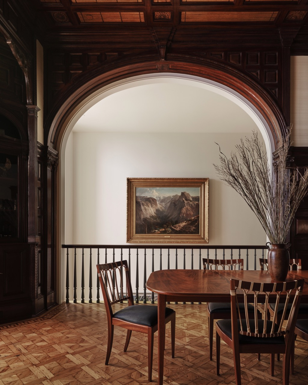 A carved wooden archway opens onto a dining area, with a landscape painting and dried branches set against the balustrade beyond