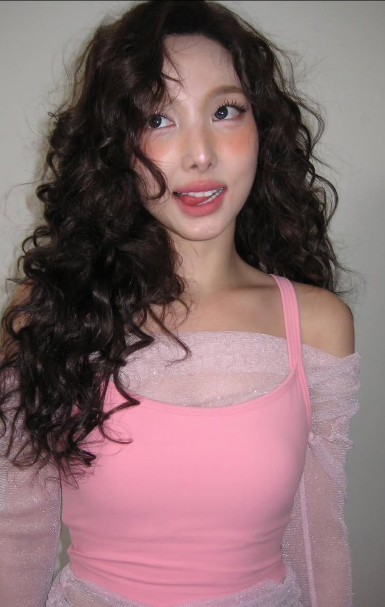 K-pop singer Nayeon wears a pink top with voluminous curls, playfully sticking her tongue out for the camera.